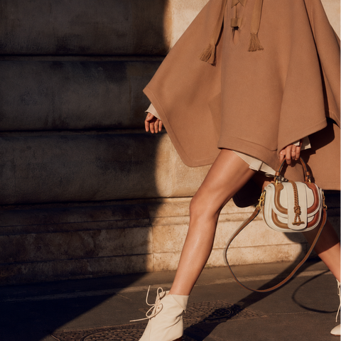 A person walking outdoors beside a stone column, wearing a camel-brown cape-style coat, white ankle boots, and carrying a cream and brown handbag with a NIKKI WILLIAMS  monogram.